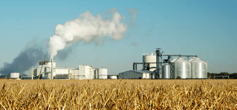 An image of a field in the foreground with an ethanol facility emitting smoke in the background.